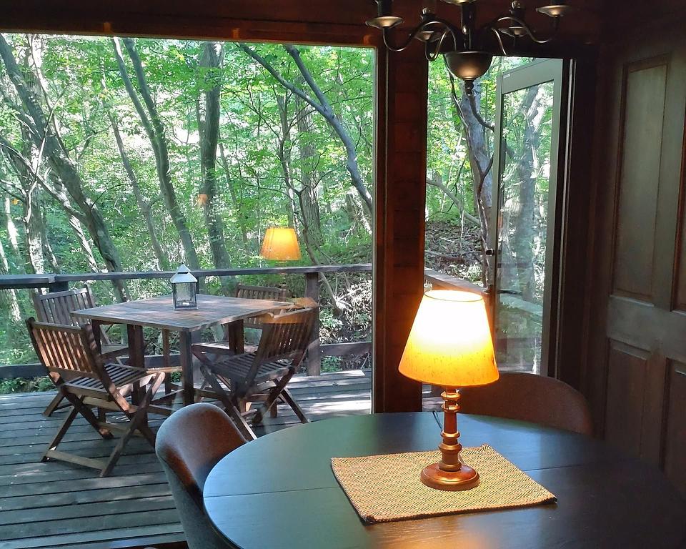 窓越しに見える緑豊かな森とウッドデッキのテーブルセットが置かれたリビングルーム。室内のテーブルには温かい光を放つランプが置かれている。