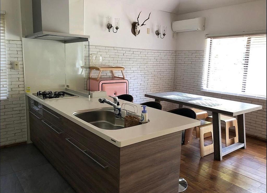 モダンなデザインのキッチンとダイニングエリア。キッチンには広々としたカウンターとシンクがあり、奥にはシンプルな木製テーブルとベンチが配置されている。白いレンガ調の壁が明るい雰囲気を演出している。